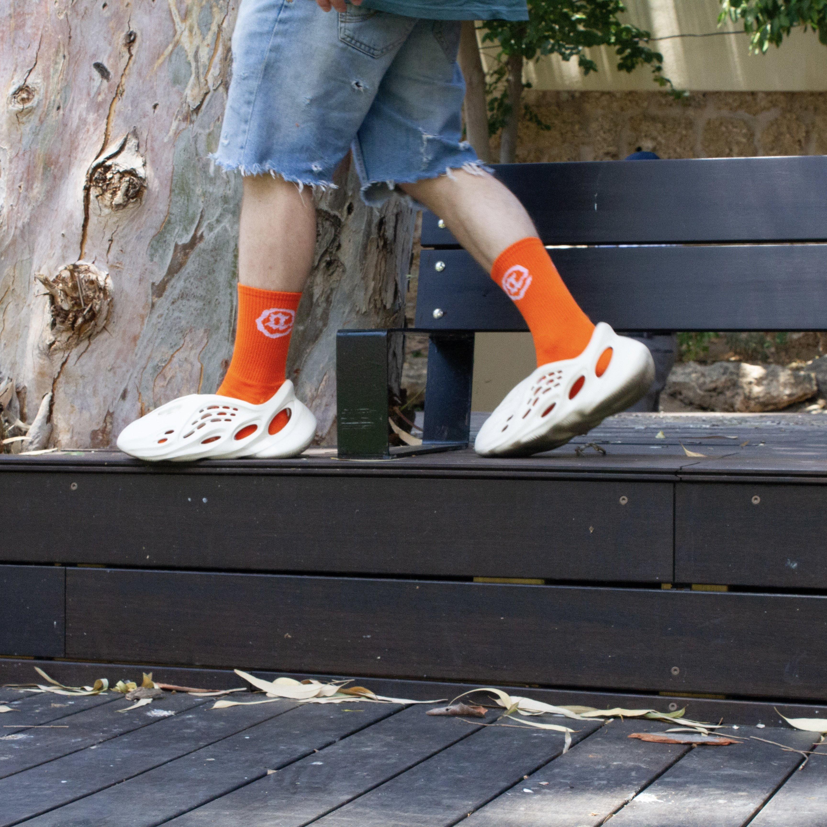 Person wearing bright orange crew socks with logo, white foam sandals, and denim shorts outdoors