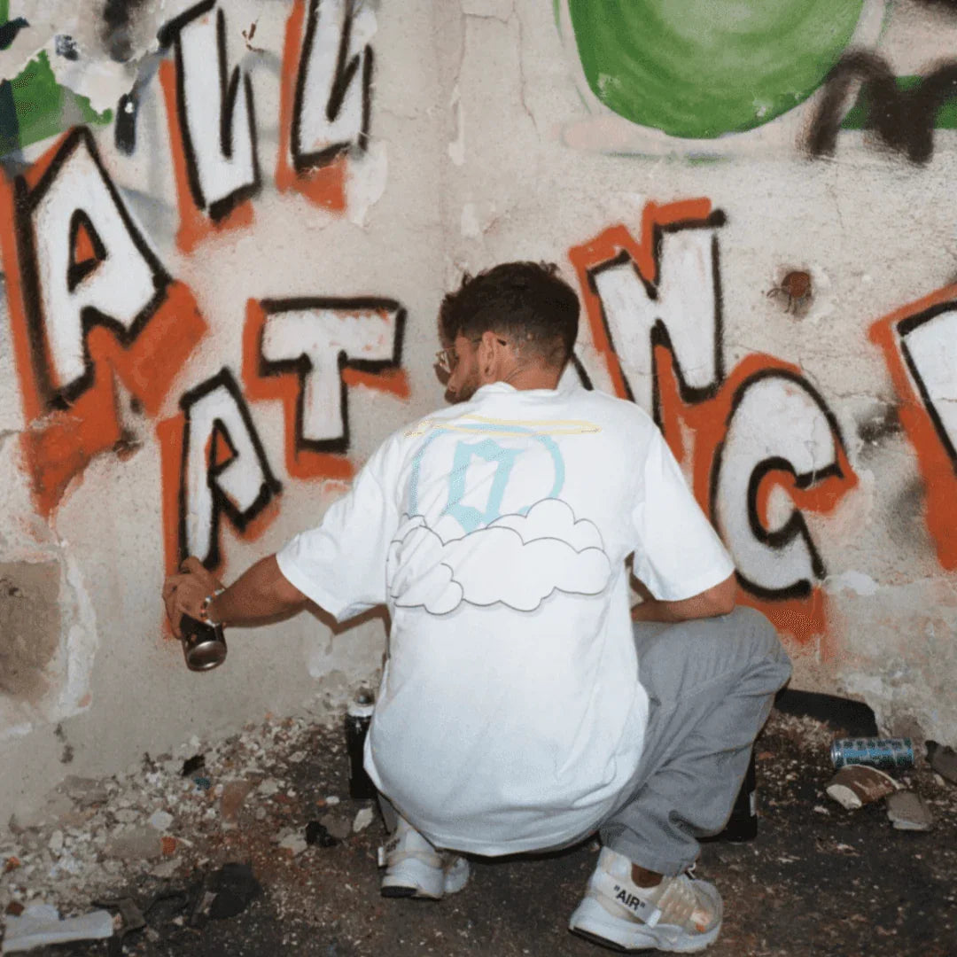 Man spray painting graffiti while wearing white heaven graphic tee with cloud and halo design