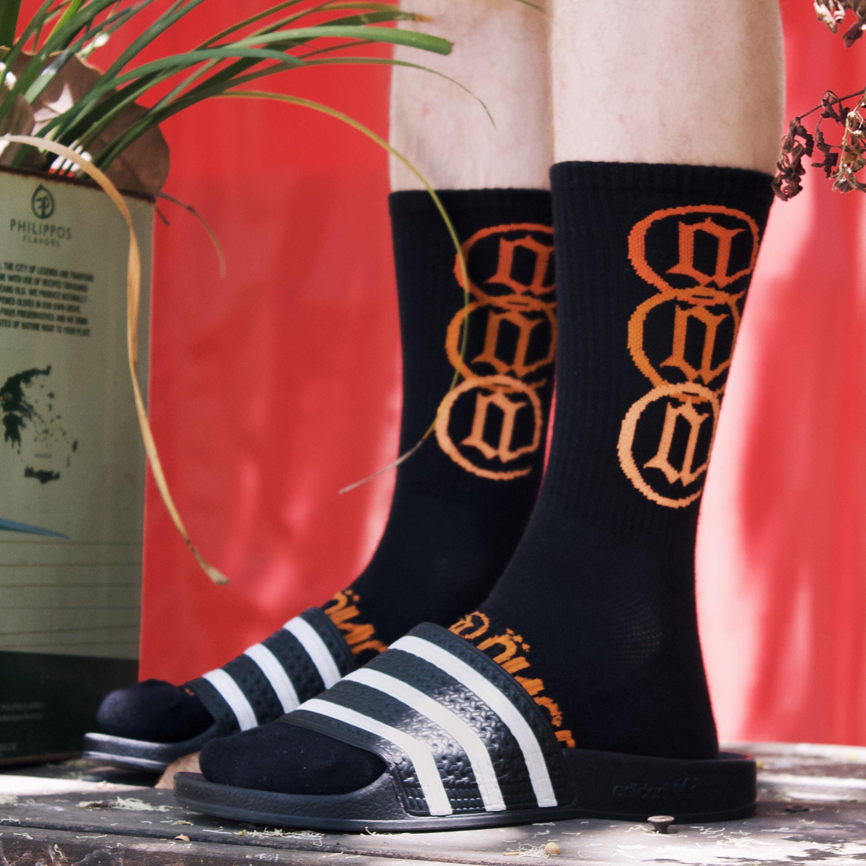 Black crew socks with bold orange graphic lettering, worn with striped slide sandals outdoors