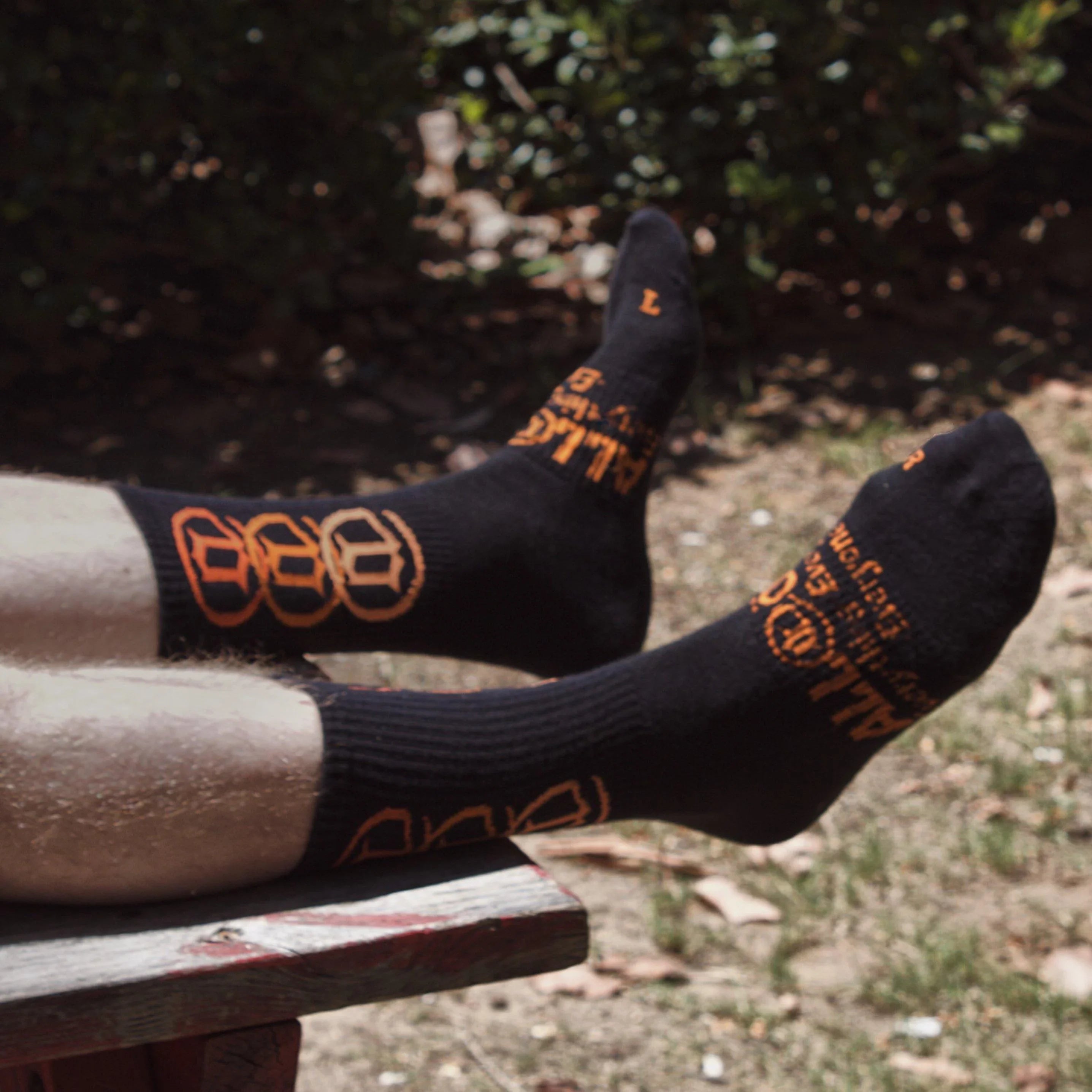 Black crew socks with orange lettering and graphic details worn outdoors on a bench