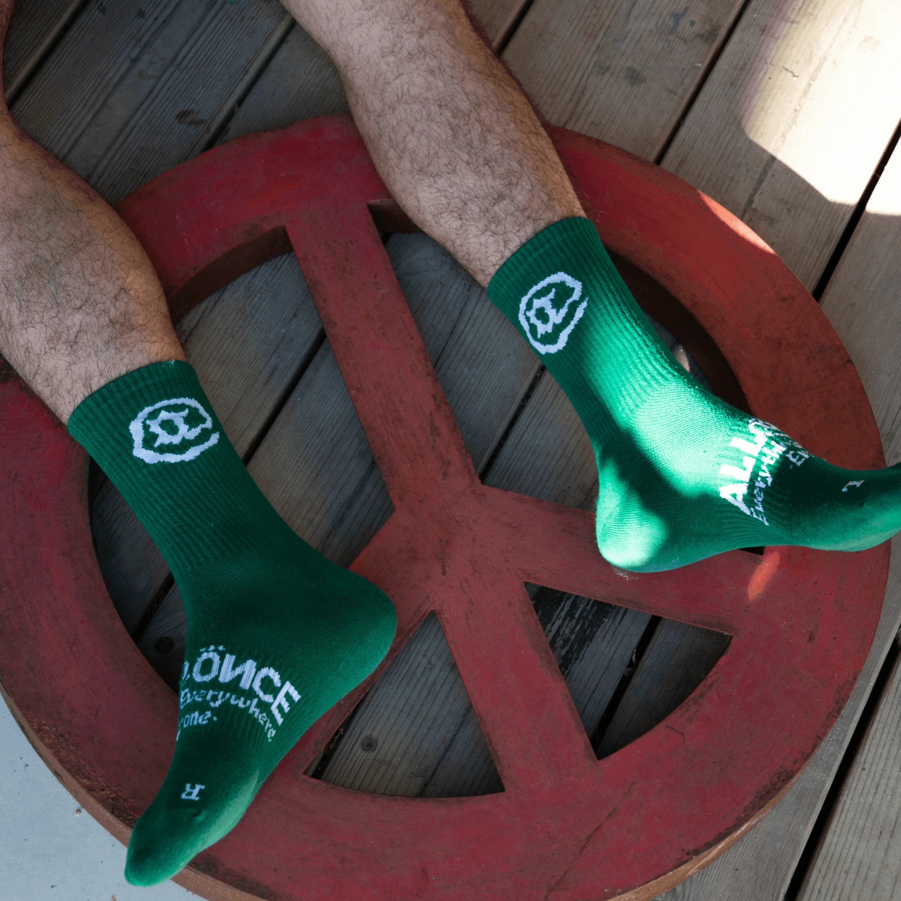 Green socks with white smiley face design and text, worn on wooden deck with peace sign