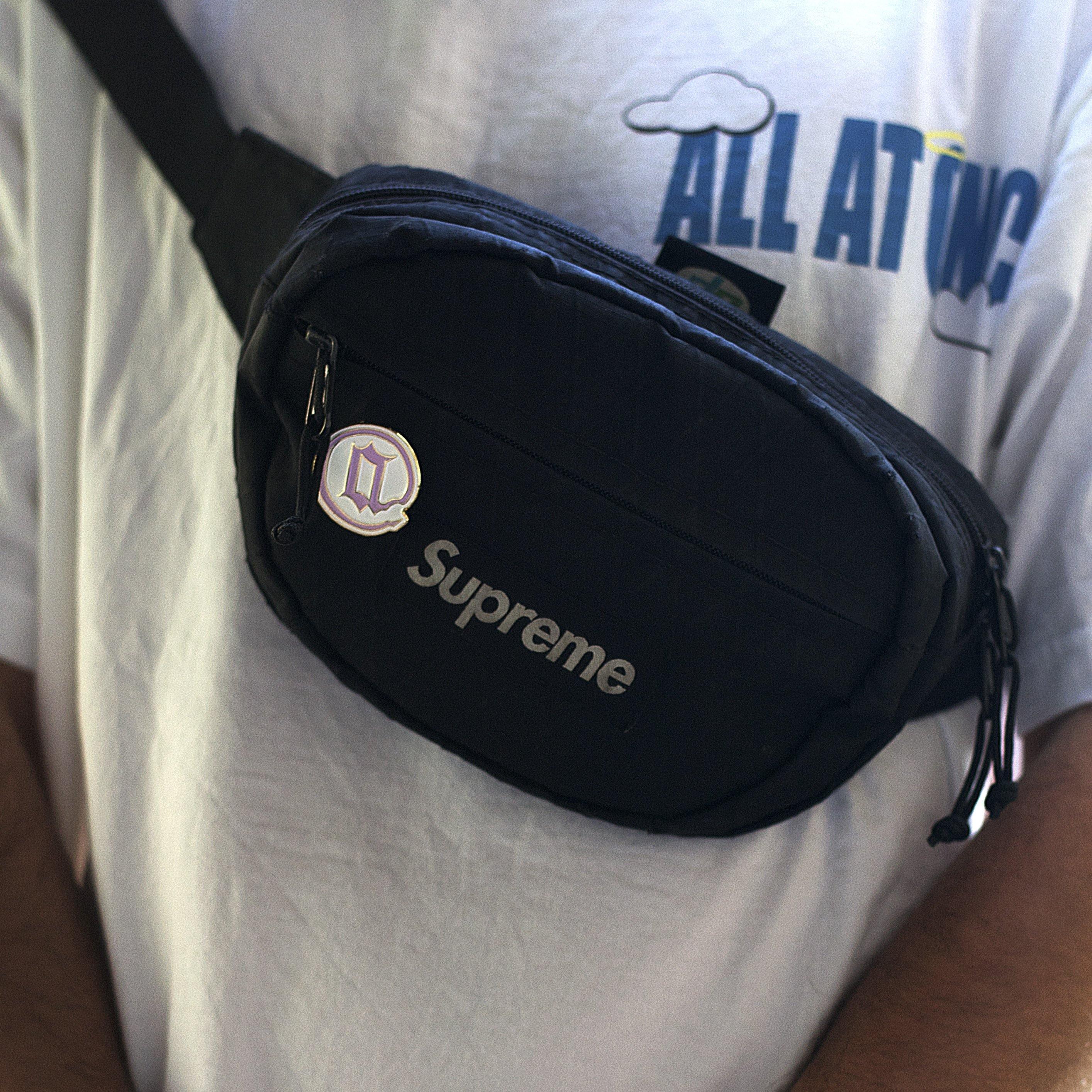 Person wearing a white graphic t-shirt and a black Supreme crossbody bag with a round enamel pin attached