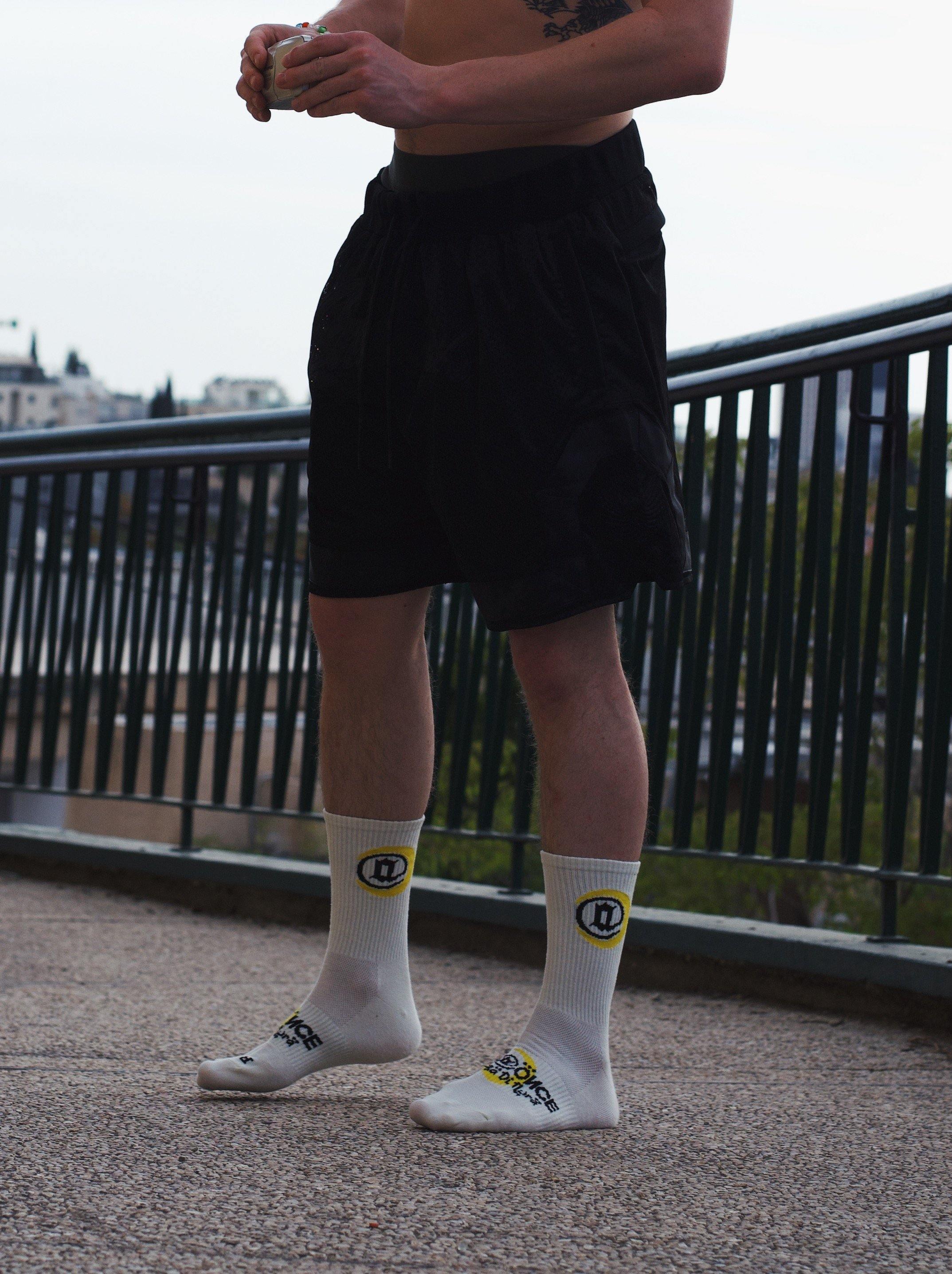 Person wearing white graphic crew socks with black shorts outdoors, yellow and black logo on socks.
