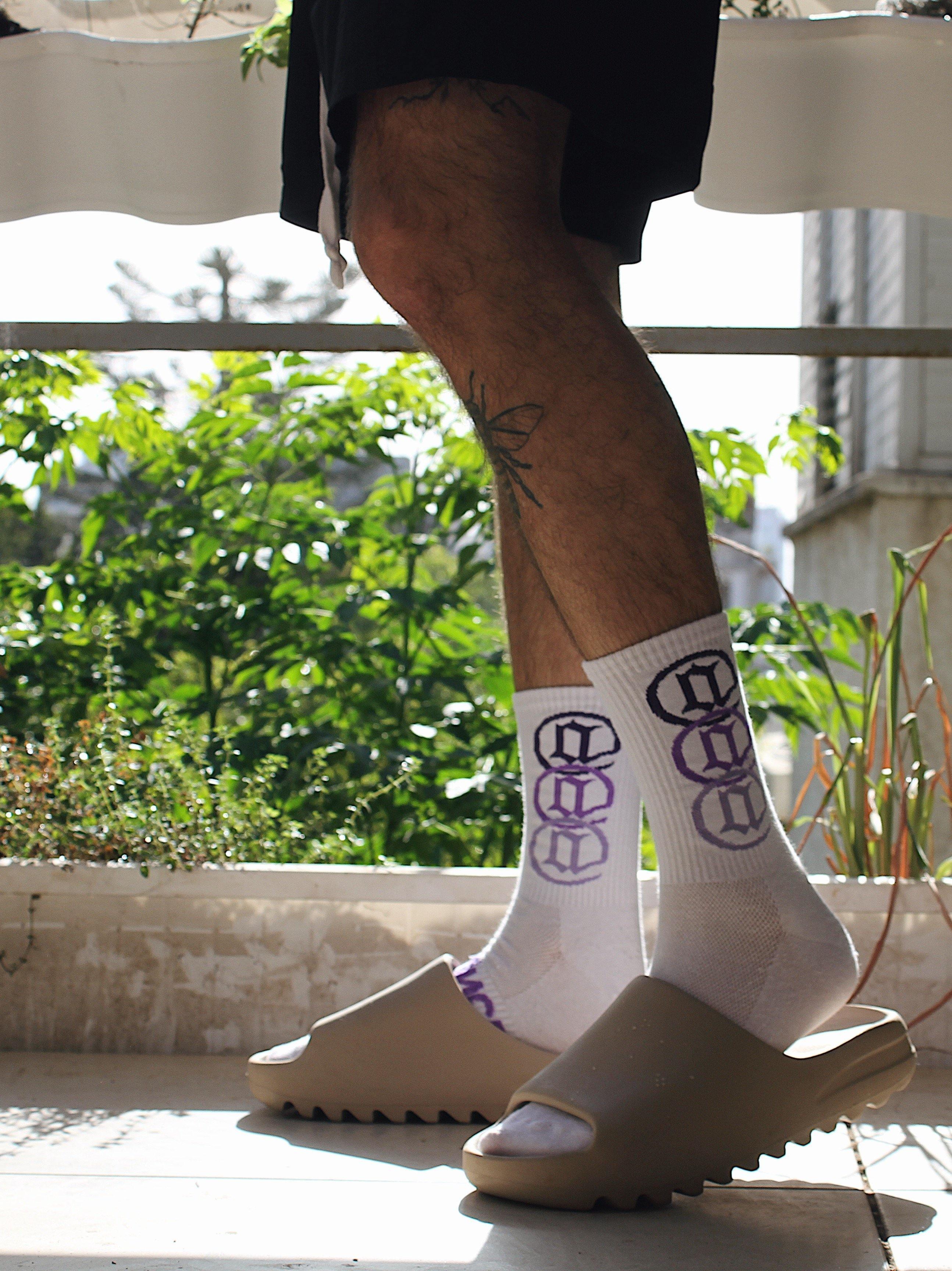 Person wearing white crew socks with purple graphic print and beige slide sandals on balcony